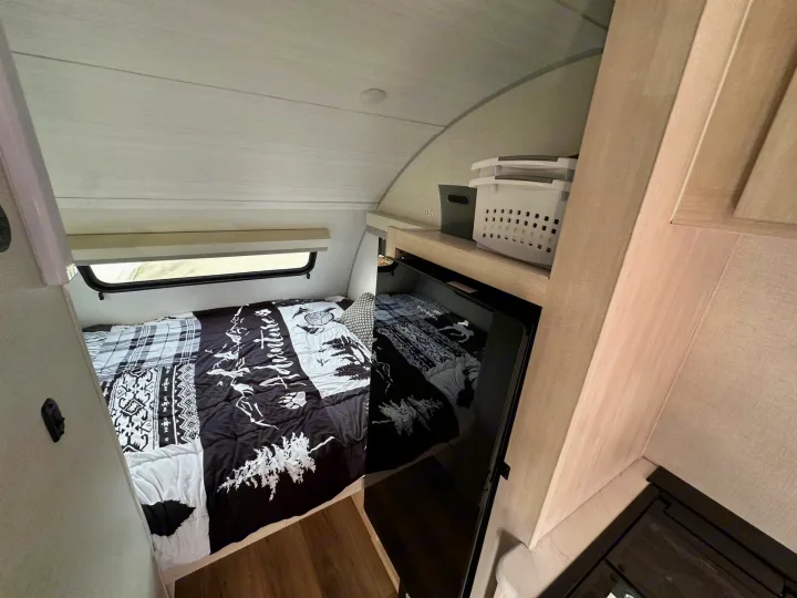 Cozy RV rental interior: features a comfortable bed with a nature-themed comforter, window, storage, and compact fridge.. Forest River RPod 2026