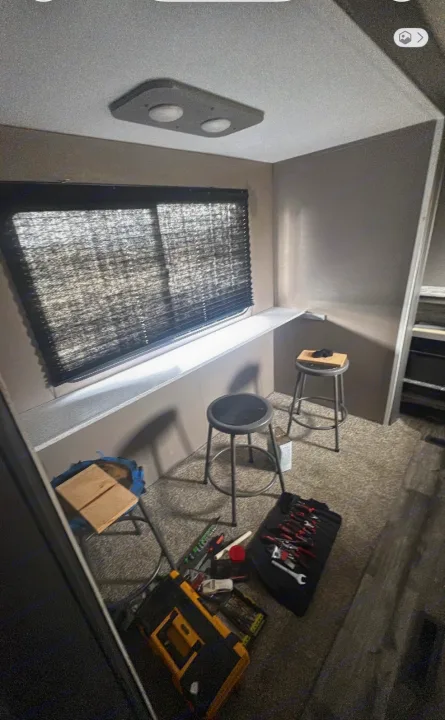 RV rental interior showing a work area with tools on carpet, two stools, a window with blinds, and a ceiling light.. Coleman Cch 2022