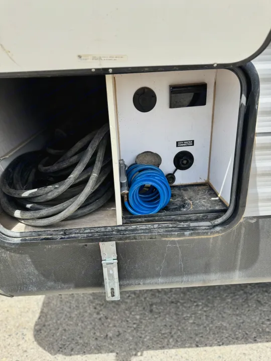 Open RV rental utility compartment with city water connection, power inlet, coiled blue hose, and electrical cord.. Coleman Cch 2022