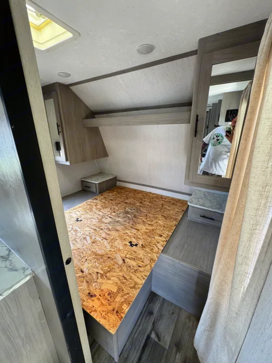 Interior of an RV rental bedroom with a wooden bed platform, overhead storage, cabinets, and a mirror reflecting the photographer.. Coleman Cch 2022