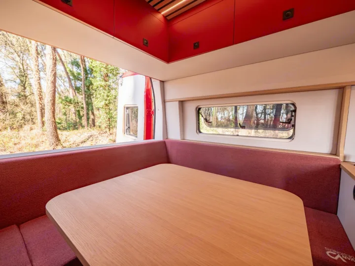 Bright RV rental interior showing a dining area with red benches and a wooden table, overlooking a serene forest through wide windows.. Ford Transit 2024