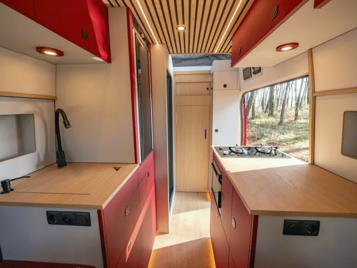 Explore this modern RV rental kitchen: black faucet, sink, stove, wood counters, and red/white cabinets, with a forest view.. Ford Transit 2024