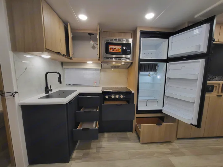 Modern rv rental kitchen featuring an open two-door fridge, microwave, stove, sink, and several open light wood/dark gray cabinets.. Winnebago Minnie Winnie 2023