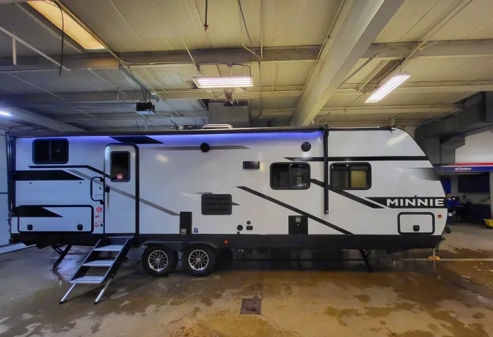 A sleek white Minnie travel trailer for RV rental, featuring blue awning lights and extended steps, is shown in a service bay.. Winnebago Minnie Winnie 2023
