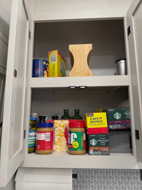 RV rental cabinet essentials: peanut butter, Wheat Thins, K-Cup coffee pods, pickles, and popcorn on shelves. Paper towels below.. Forest River Coachman Chaparral 2025