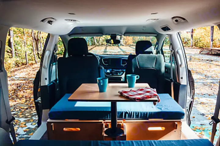 Enjoy fall camping! This RV rental offers a converted minivan interior with a table, mugs, and comfy seating setup.. Dodge 2022
