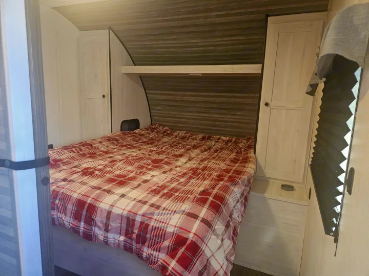 RV rental bedroom with a comfy plaid bed, built-in side cabinets, an overhead shelf, and pleated window shade.. Heartland RVs Trail Runner 2024