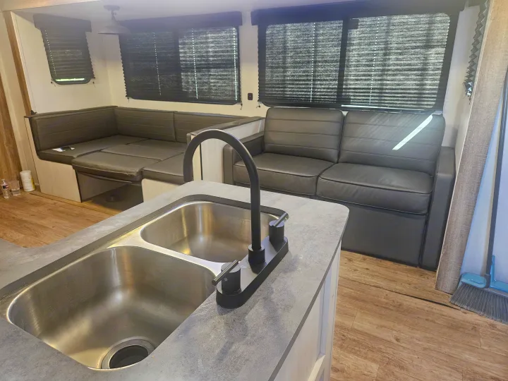 Step inside this RV rental's modern interior, showcasing a dual kitchen sink, sleek black faucet, and dark gray seating with window blinds.. Heartland RVs Trail Runner 2024