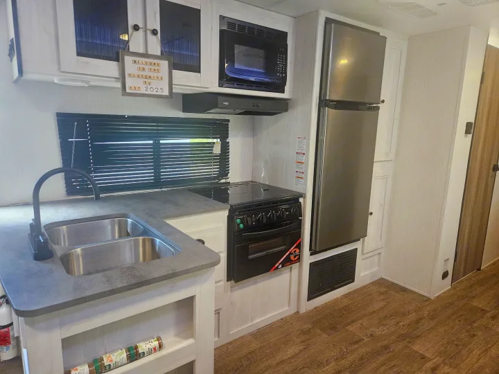 Inside an rv rental kitchen with a fridge, microwave, stove, dual sink, and "Kluczniks RV" sign.. Heartland RVs Trail Runner 2024