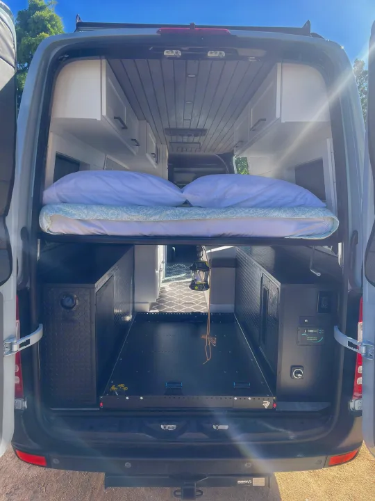 Inside a modern rv rental camper van, featuring an elevated bed, pull-out cargo tray, custom cabinets, and essential controls.. Mercedes-Benz Sprinter RV Motorhome Campervan 2012