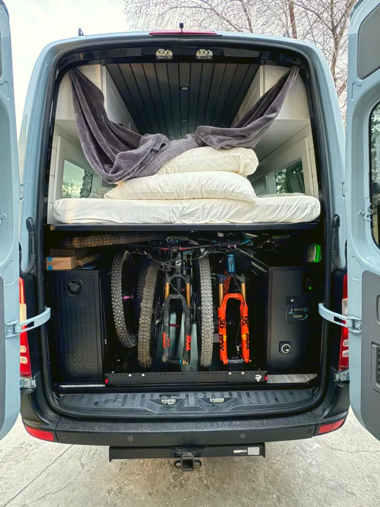 This adventure-ready rv rental shows a bed, hammock, and two mountain bikes stored in the lower garage of a grey camper van.. Mercedes-Benz Sprinter RV Motorhome Campervan 2012