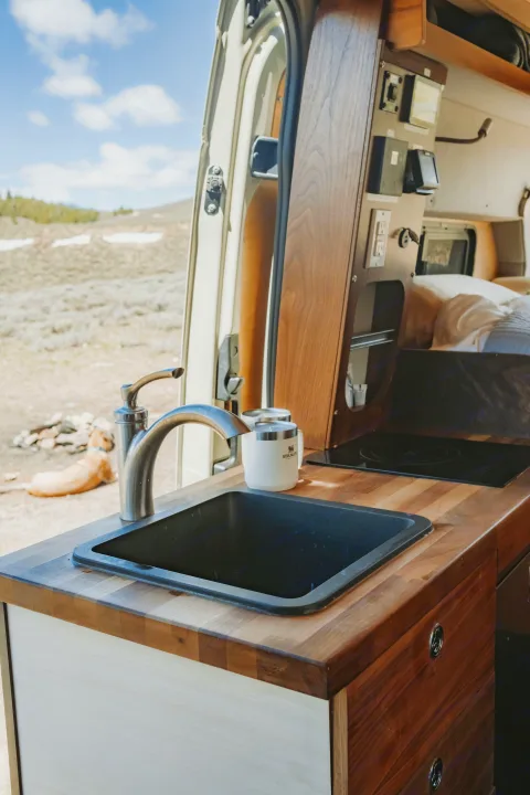 Inside a cozy RV rental: sleek kitchen with sink, stovetop, and custom wood finishes, overlooking a scenic outdoor landscape.. Custom Mercedes Benz Sprinter 2022
