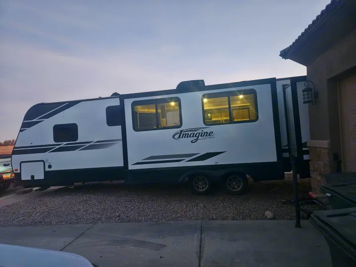 Spacious RV rental: a white travel trailer with a slide-out extended and interior lights on, parked on gravel next to a house.. Imagine Imagine design 2020