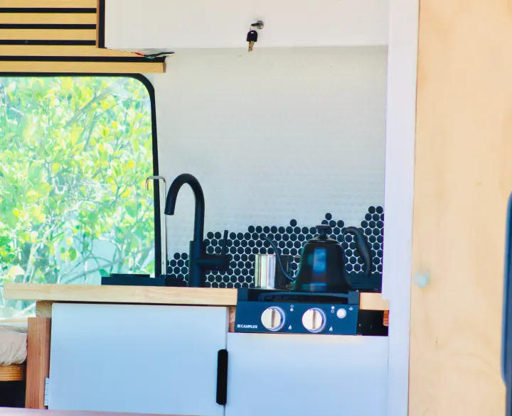 An RV rental's cozy kitchen features a black faucet, penny tile backsplash, stovetop with a kettle, and a leafy view outside.. Mercedes-Benz Sprinter 2008