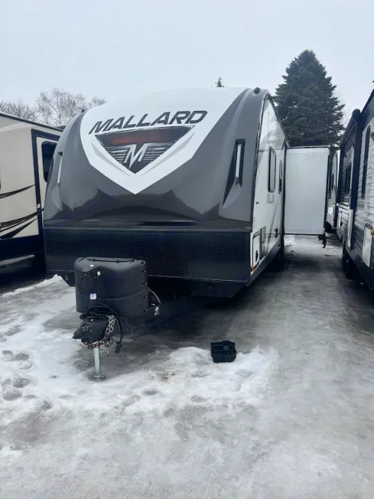 A grey and white Mallard travel trailer for RV rental, parked on snow. Visible front hitch and propane tanks.. Heartland RVs Mallard 2019