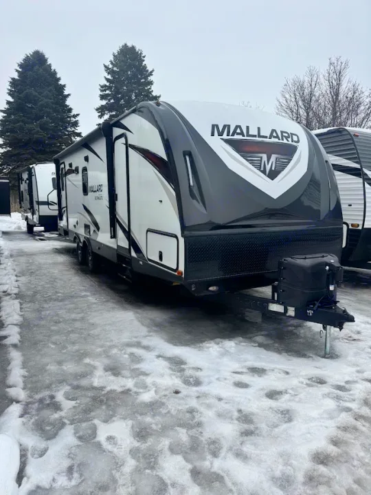 Mallard travel trailer RV rental, white and black, parked on an icy lot. Snow-covered ground with other RVs and trees.. Heartland RVs Mallard 2019
