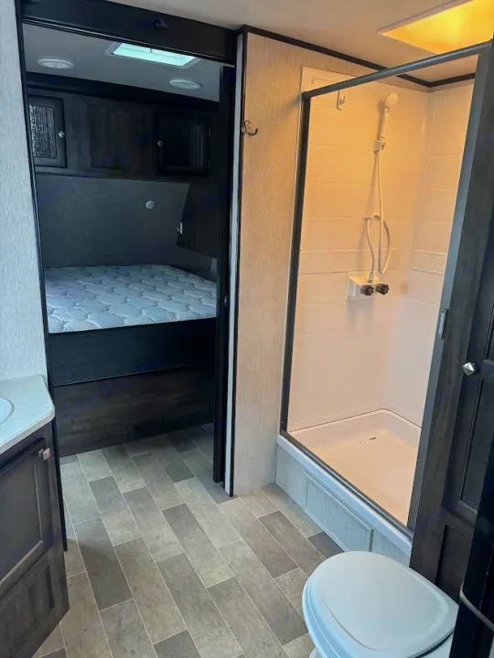 Inside an RV rental: a bedroom with a comfortable bed, adjacent to a compact bathroom with a shower, toilet, and sink.. Heartland RVs Mallard 2019