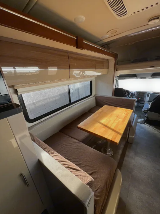 Bright RV rental interior: wooden dinette table, brown bench seating, overhead cabinets, and view of the driver's cabin.. Winnebago View 2016