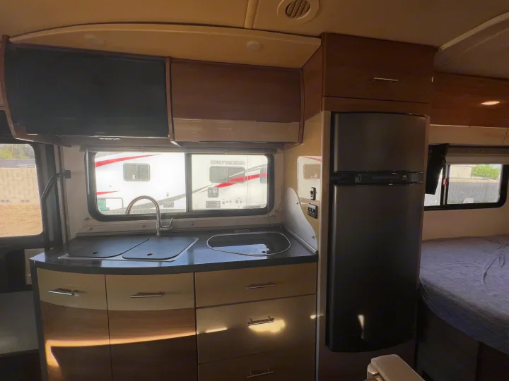 This rv rental features a cozy interior with a kitchen area, including a sink, fridge, and an overhead TV, alongside a bed.. Winnebago View 2016
