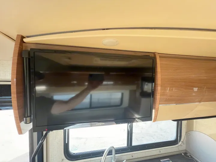 RV rental interior with a mounted TV, overhead cabinet, and counter. A person's arm reflects on the TV screen.. Winnebago View 2016