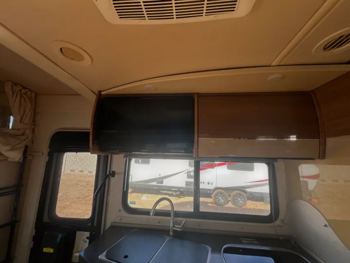 Inside an RV rental: kitchen counter with sink & faucet, overhead TV & cabinets. Windows show another RV parked outside.. Winnebago View 2016
