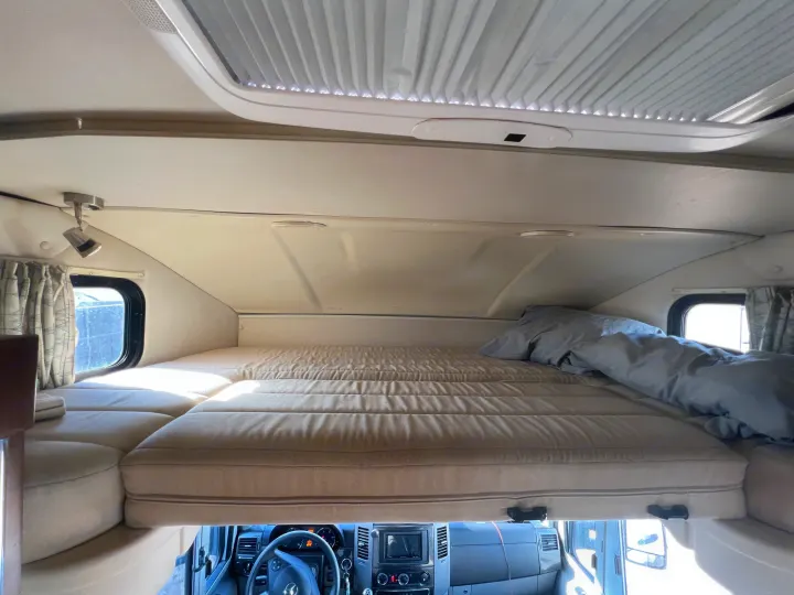 Overhead bunk bed in an RV rental, showing the comfy sleeping area above the driver's cabin with a window and ceiling vent.. Winnebago View 2016