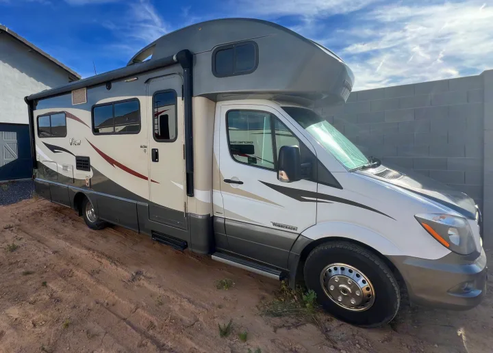 This white and gray Class C Winnebago View rv rental is parked on dirt, ready for your next adventure under a blue sky.. Winnebago View 2016