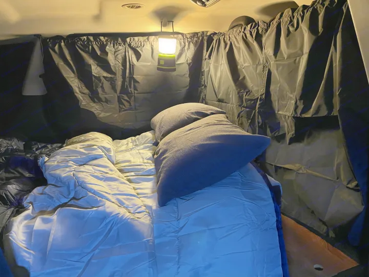 Cozy RV rental car camping setup featuring a bed with pillows, privacy curtains, and a bright lantern for comfortable nights.. Dodge Journey 2017