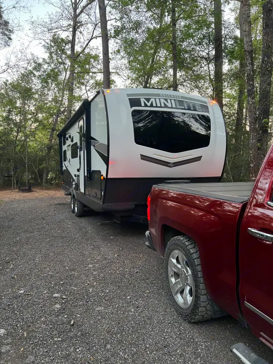 Red pickup truck hitched to a white Rockwood Mini Lite travel trailer on a gravel campsite among trees. Your ideal RV rental setup.. Forest River Rockwood Mini Lite 2024
