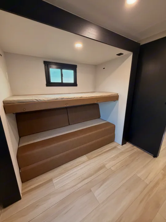 Modern RV rental bedroom/lounge: features an upper bunk bed over a convertible brown sofa, window, and light wood floor.. Forest River Salem Hemisphere 2026