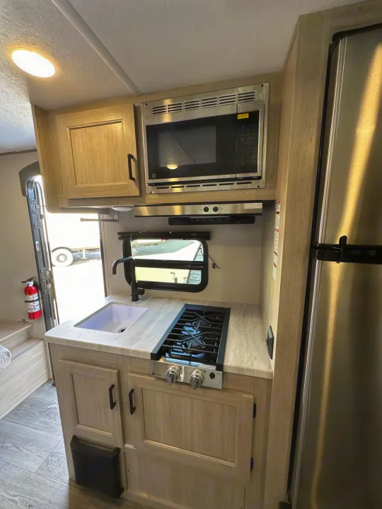 RV rental kitchen: light wood cabinets, microwave, stove, sink, refrigerator, and an open door with a fire extinguisher.. Coachmen CATALINA 2025