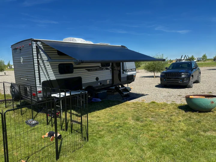 RV rental setup: A modern travel trailer with awning, black Jeep SUV, pet playpen, and fire pit under a clear blue sky. Camping ready!. Coachmen CATALINA 2025