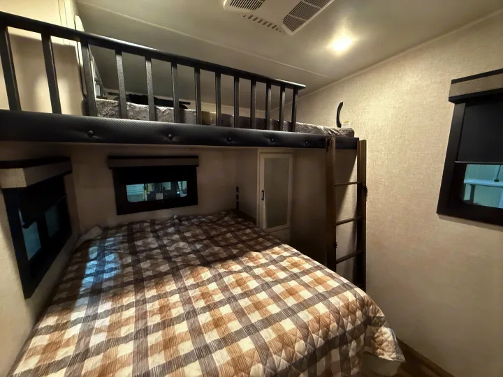 RV rental bedroom with a plaid lower bed, upper bunk bed with safety railing, ladder, and windows. Cozy mobile sleeping area.. Jayco Eagle 2025