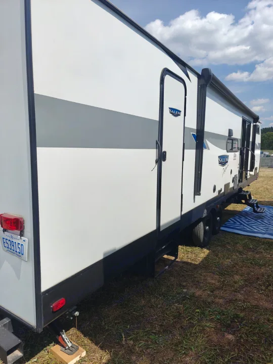 This rv rental is a white travel trailer with grey accents, doors, and steps, parked on grass under a cloudy sky. A blue mat is outside.. Forest River Salem 2024