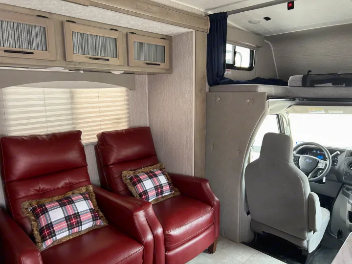 Cozy RV rental interior with red recliners, plaid pillows, overhead cabinets, and a bunk bed over the driver's cabin.. Coachmen Freelander 2021