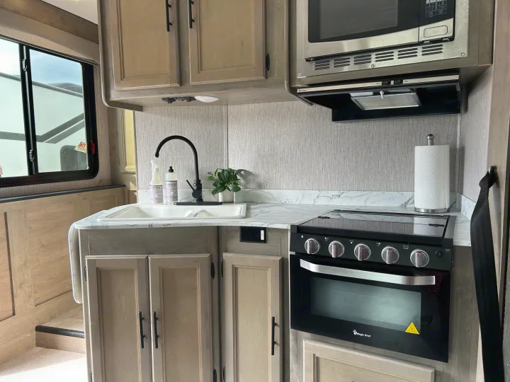 Modern RV rental kitchen with light wood cabinets, featuring a sink, microwave, and Magic Chef stove for your travel cooking needs.. Coachmen Freelander 2021