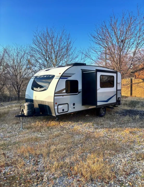 A modern white & grey Sonic travel trailer, parked on dry grass with bare trees in background, available for RV rental.. Sonic Lite 2021