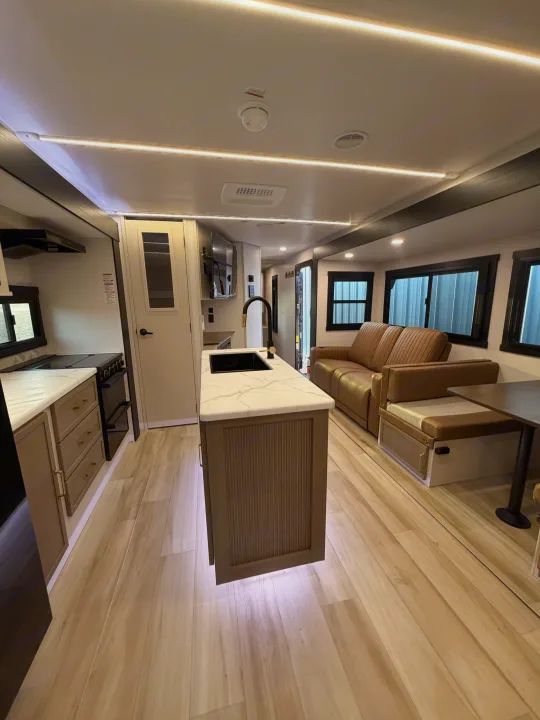 Modern RV rental interior: bright kitchen island, brown couch, light wood floors, and ceiling LED strip lighting.. Forest River Salem Hemisphere 2026