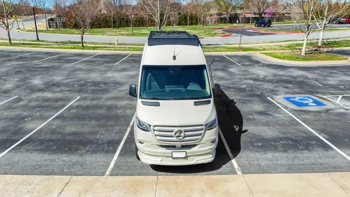 White Mercedes-Benz Sprinter rv rental, with roof rack, parked in lot next to handicapped space. Bare trees in background.. OGV Luxury Coach V-Cruise 2026