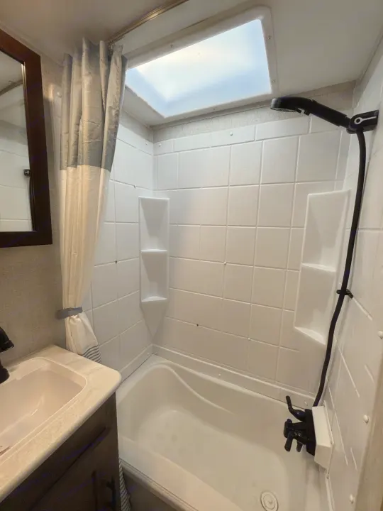 Inside an RV rental bathroom: a tub/shower with skylight, black showerhead, corner shelves, sink, and mirror.. Forest River Wildwood 2020