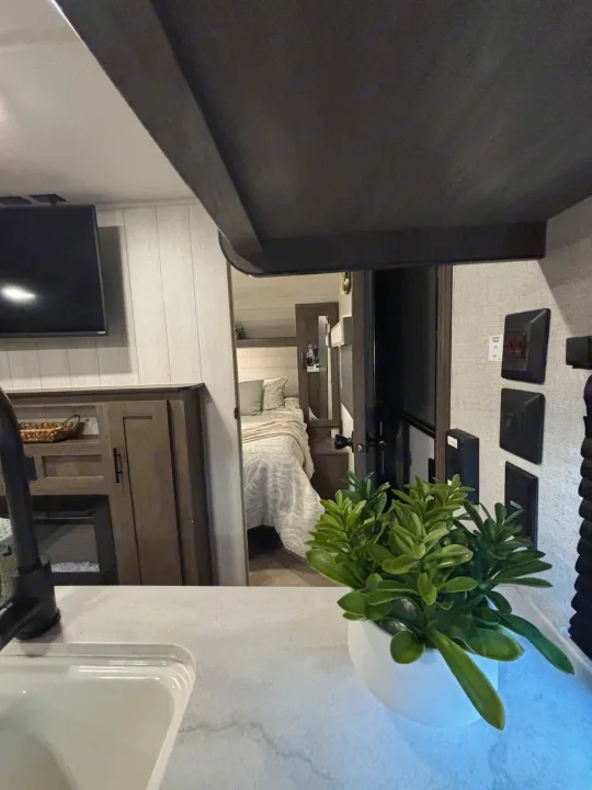 Modern RV rental interior: kitchen counter with a faucet, a green plant, and a view into the cozy bedroom.. Forest River Wildwood 2020