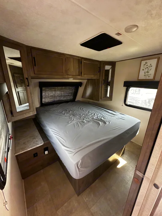 This RV rental bedroom features a grey bed, overhead wooden cabinets, a side window, and a built-in nightstand.. Coachmen Apex 2021