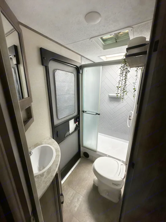 Explore this clean RV rental bathroom, offering a compact sink, toilet, and shower with modern gray herringbone tiles and a skylight.. Coachmen Apex 2021