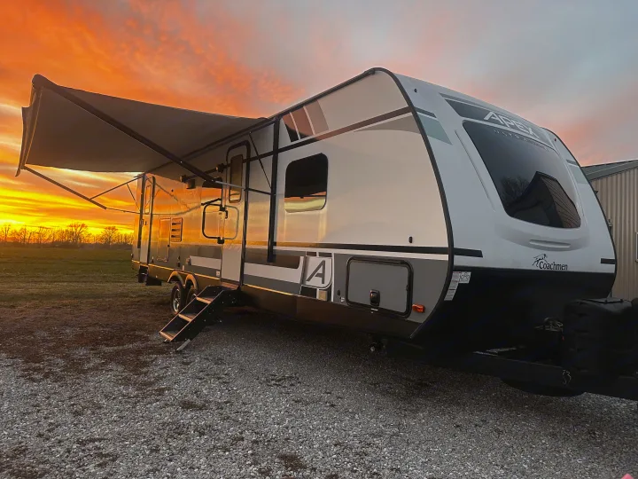 RV rental travel trailer with awning extended, glowing under a vibrant orange and yellow sunset by a field.. Coachmen Apex 2021
