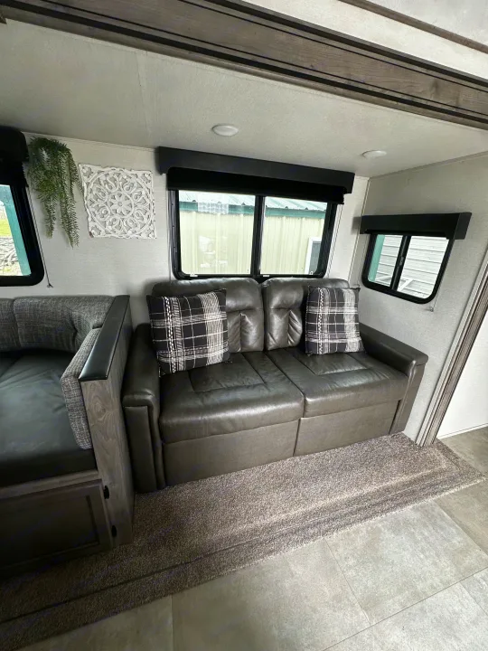 Cozy RV rental interior with a dark gray leather sofa, plaid pillows, and a soft gray rug. Light walls and windows with blinds.. Coachmen Apex 2021