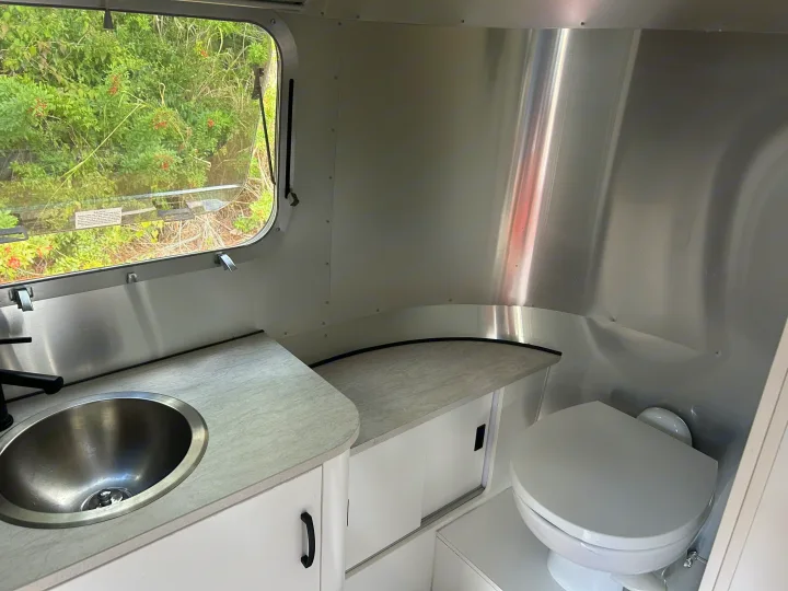 RV rental interior: A compact wet bath featuring a stainless steel sink, modern black faucet, toilet, and bright window to nature.. Airstream Caravel 2022
