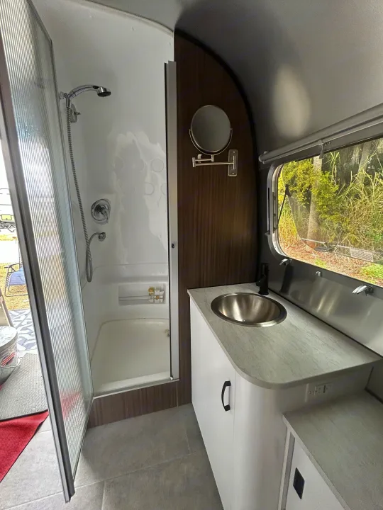 Compact RV rental bathroom with shower, stainless steel sink vanity, and window views. Ideal for travel trailer adventures.. Airstream Caravel 2022