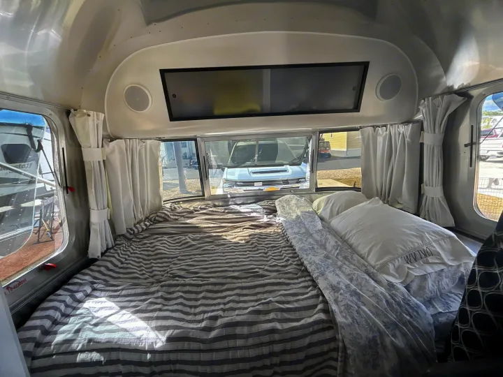 Interior of an Airstream **rv rental**: a comfy bed with striped bedding, white curtains, and windows offering an outdoor view.. Airstream Caravel 2022