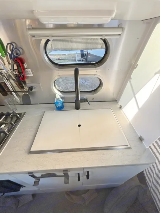 RV rental kitchen: modern sink, black faucet, stovetop, and oval windows looking outside. Clean and functional interior.. Airstream Caravel 2022
