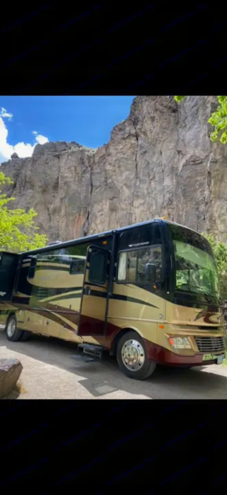 This detailed RV rental, a black, tan, and burgundy motorhome with extended slide-outs, rests before a towering rock cliff.. Fleetwood Bounder 2013